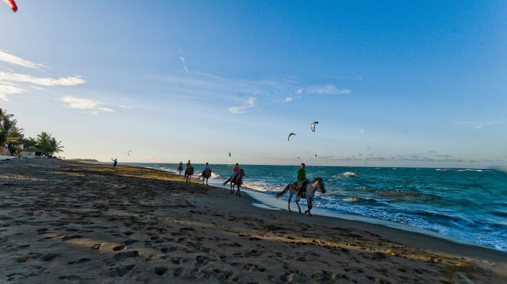 people-horseback-riding-on-beach-sand-turquoise-ocean-sunset