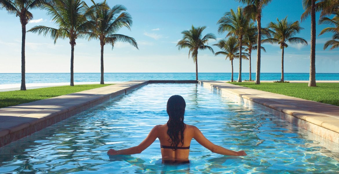 back-of-woman-in-pool-overlooking-ocean-green-palm-trees