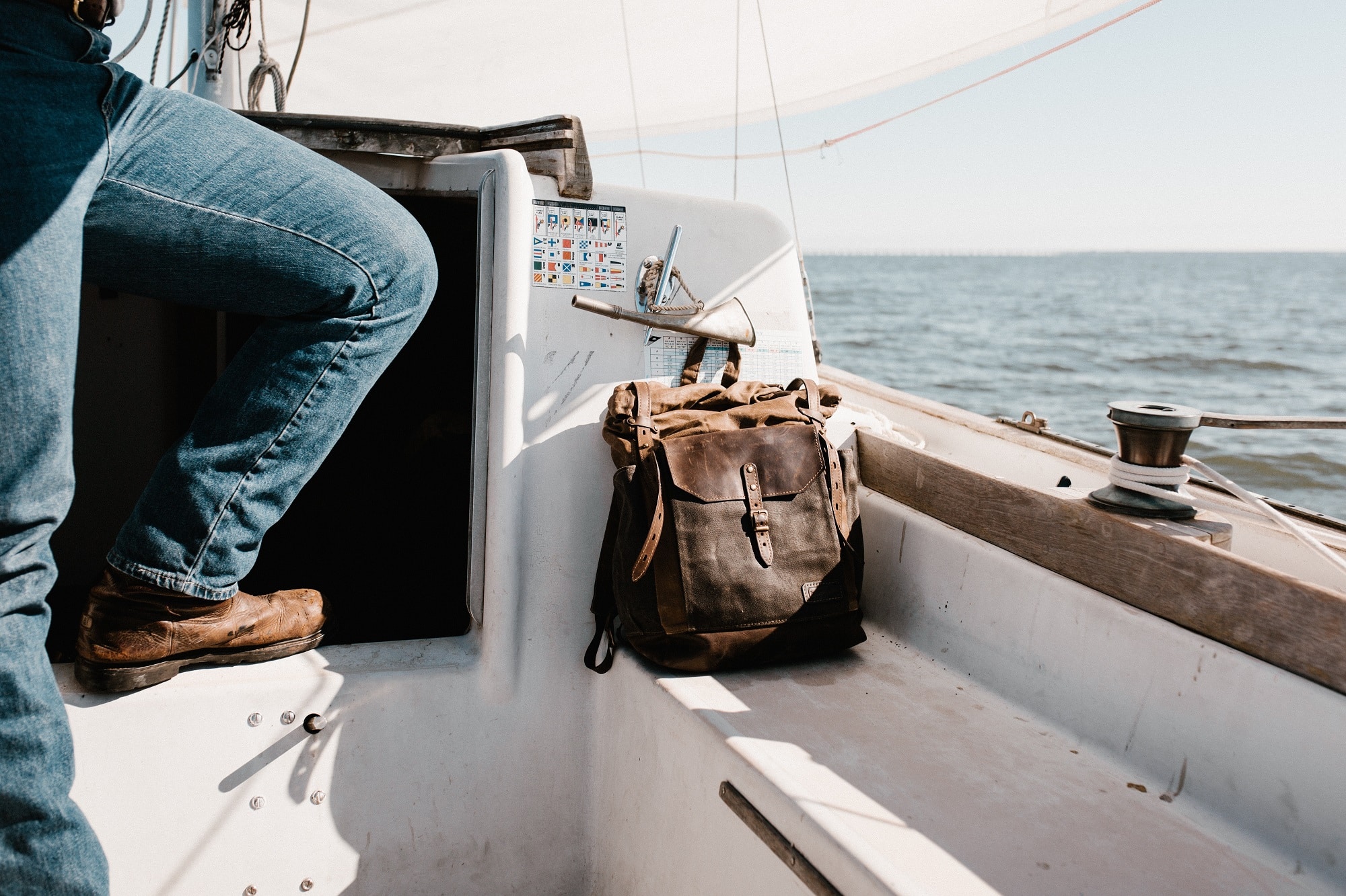 Someone wearing jeans and boots perches their leg and backpack upon the deck of a sailboat with the water in the background.
