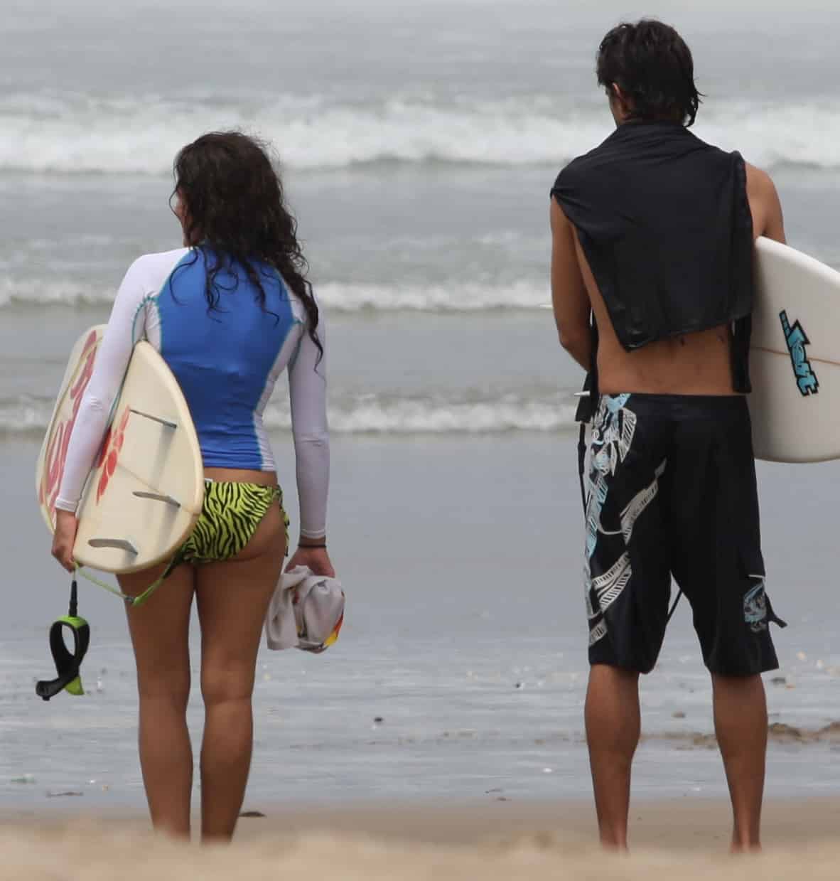 Two surfers stand facing the water, one wears a rashguard longsleeved shirt