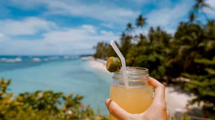 Someone holds an alcoholic beverage in a mason jar up with the backdrop of a beautiful natural beach