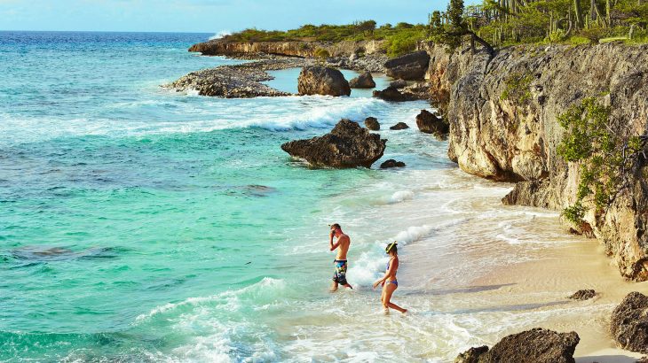 Couple with snorkel mask and gear walking into turquoise ocean on secluded beach with cliffs in the background