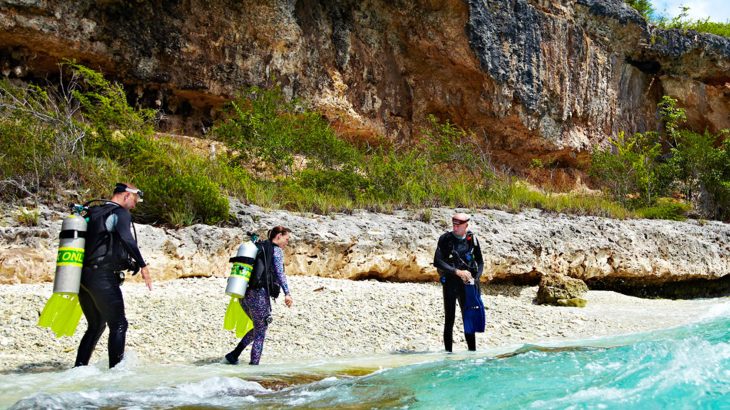 Three divers with oxygen tanks walking into blue water from a beach with huge cliffs behind them