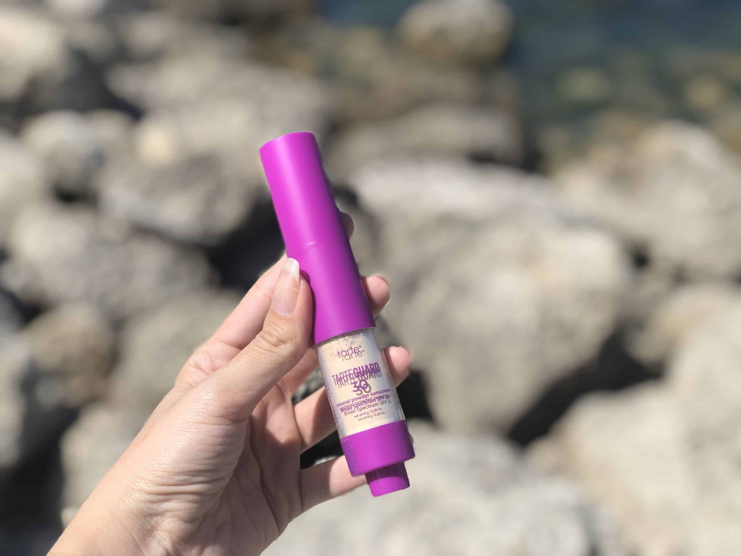 A hand holds a container of Tarte Tarteguard SPF 30 powder sunscreen in front of rocks at the beach.