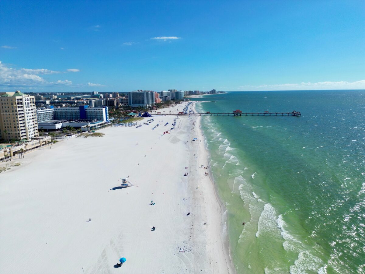 Aerial view of Clearwater Beach, Florida, featuring white sands, turquoise waters, and a distant pier under a clear blue sky