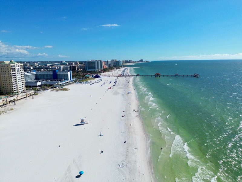 Aerial view of Clearwater Beach, Florida, featuring white sands, turquoise waters, and a distant pier under a clear blue sky