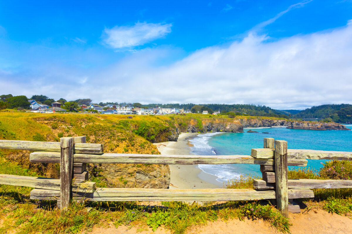 View through rustic wooden fence of sand beach below Main Street and houses of Mendocino town communinty on a sunny summer day in California