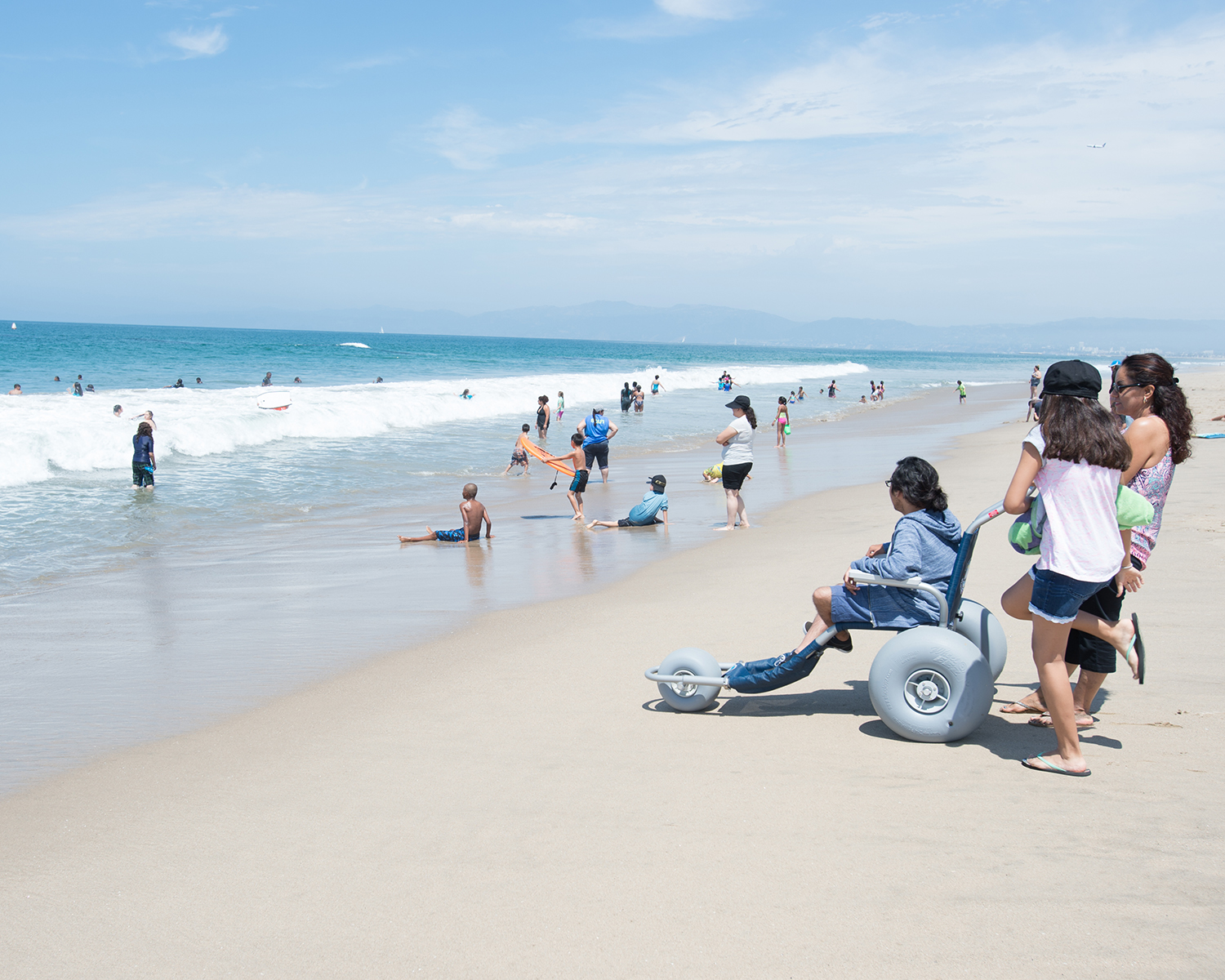 A person in a balloon-tire beach wheelchair at the Dockweiler beach shore at midday.